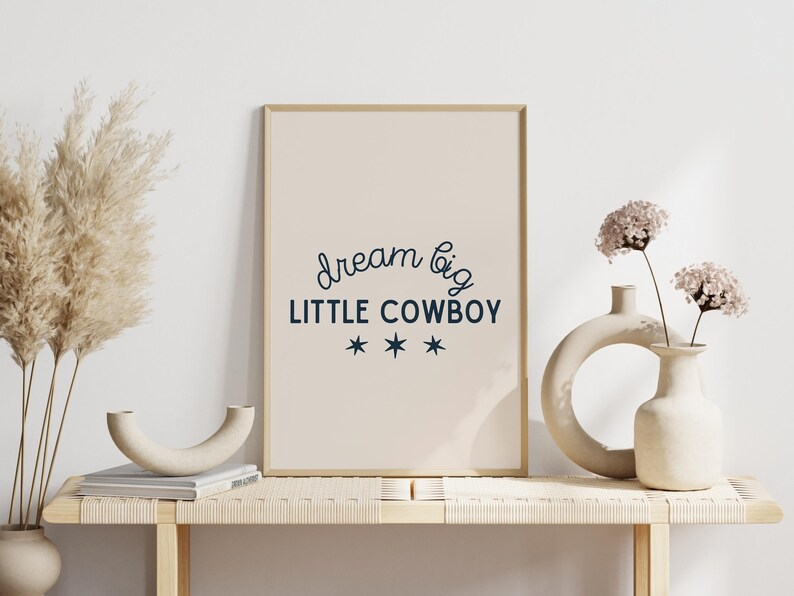 May include: A framed print with a beige background and the text "dream big LITTLE COWBOY" in dark blue with three stars underneath. The print is sitting on a light wood shelf with two white ceramic vases and dried pampas grass.