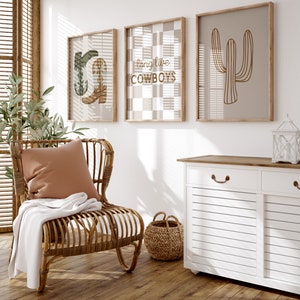 May include: A white cabinet with two drawers and two doors, a wicker chair with a brown throw blanket and a pillow, a wicker basket, and three framed prints with a cactus, cowboy boots, and a checkered pattern with the text "long live cowboys".