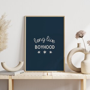 May include: A navy blue framed print with white text that reads "long live boyhood" with three white stars underneath. The print is sitting on a light wood shelf with two white ceramic vases and dried pampas grass.