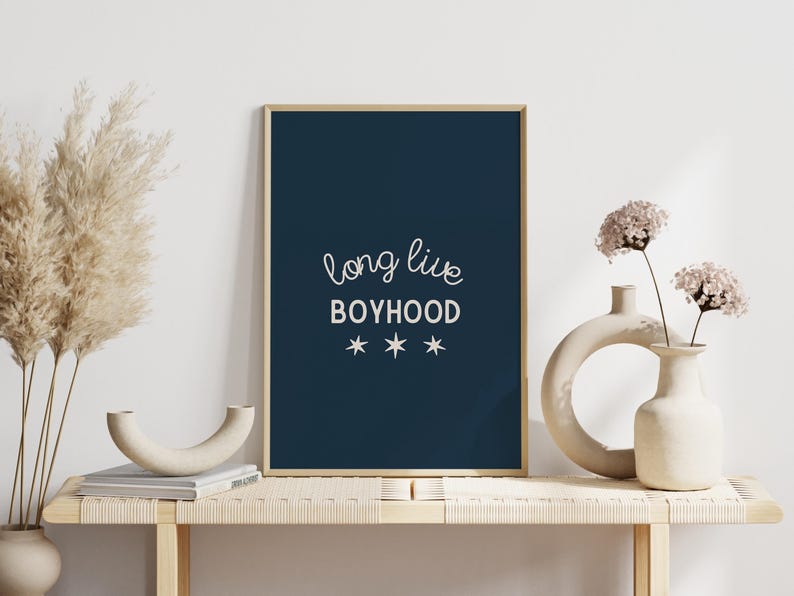 May include: A navy blue framed print with white text that reads "long live boyhood" with three white stars underneath. The print is sitting on a light wood shelf with two white ceramic vases and dried pampas grass.