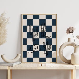 May include: A framed print with a black and white checkered background. The print features a cowboy hat, cactus, and cowboy boots in black outlines.
