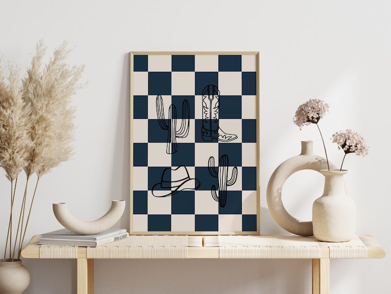 May include: A framed print with a black and white checkered background. The print features a cowboy hat, cactus, and cowboy boots in black outlines.