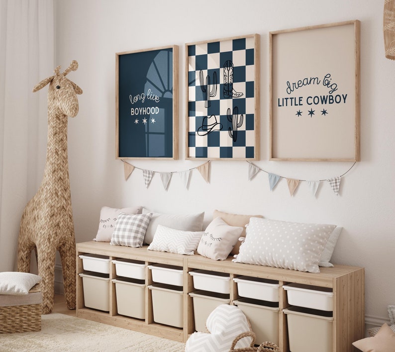 May include: A nursery room with a light blue, white, and brown color scheme. There are three framed prints on the wall with the text "long live boyhood", a checkered pattern with cacti and cowboy boots, and "dream big little cowboy". There is a wooden bench with storage bins and pillows. A stuffed giraffe stands next to the bench.
