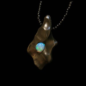 May include: A dark brown pendant with an irregular shape, featuring a round, iridescent opal stone. The pendant hangs from a dark chain necklace against a black background. The opal displays shades of blue, green, and white.