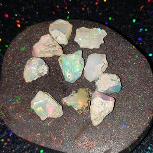 May include: A collection of eight rough opal stones, with a variety of shapes and sizes, displayed on a dark gray background. The opals have a milky white base with flashes of iridescent color.