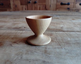 Puede incluir: Un huevero de madera con un cuenco redondeado y una base acampanada. El huevero está hecho de madera marrón claro y tiene un acabado liso.