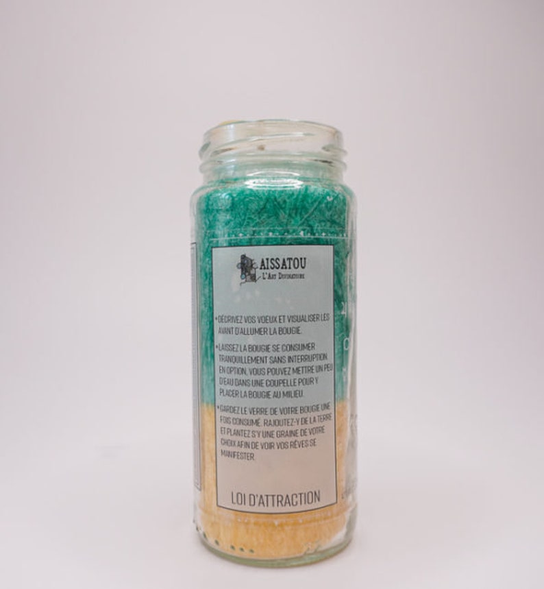 May include: A glass jar candle with a green and yellow wax blend. The label reads "AISSATOU L'Art Divinatoire" and "LOI D'ATTRACTION". The candle instructions are in French.