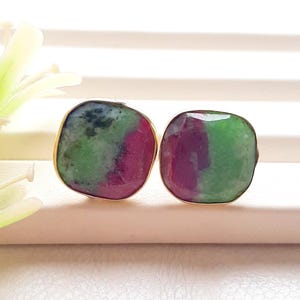 May include: A pair of stud earrings featuring square-shaped, faceted ruby zoisite stones with a polished finish. Each stone displays a natural blend of vibrant green and deep magenta-pink hues, encased in a thin, simple gold-toned metal setting.