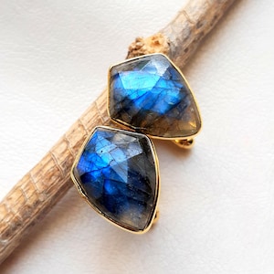 May include: A pair of gold-plated stud earrings with faceted labradorite stones. The stones have a shimmering blue and grey appearance, set in a geometric shape. The earrings are presented on a light background.