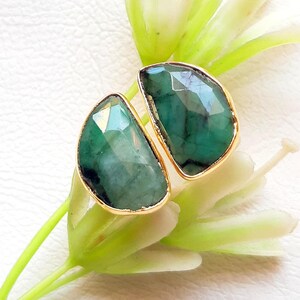 May include: Two emerald-green gemstone rings with gold settings. The faceted gemstones are displayed on a white surface with green and white floral accents. The rings are a statement piece.