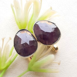 May include: A pair of cufflinks featuring large, faceted, deep purple, oval-shaped gemstones set in gold-toned metal frames. The stones have a polished, reflective surface with visible geometric facets, and the metal settings are smooth and rounded.