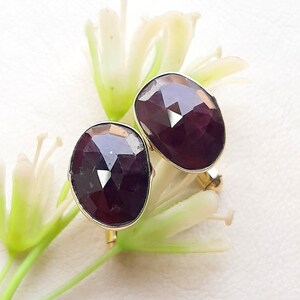 May include: A pair of oval-shaped cufflinks featuring deep purple, faceted gemstones set in gold-toned metal. Each cufflink has a polished, multi-faceted surface that reflects light, showcasing a dark, rich hue and a smooth, rounded edge design.
