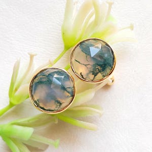 Puede incluir: Un par de pendientes de tono dorado con piedras de ágata verde y blanca. Las piedras son redondas y tienen un diseño facetado.