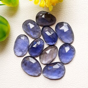 Peut inclure: Dix pierres précieuses facettées en forme de larme, de couleur bleu-violet. Les pierres sont disposées en grappe sur une surface blanche.