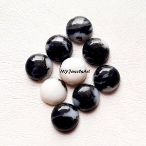 Puede incluir: Una colección de nueve cabujones redondos y pulidos que presentan una mezcla de patrones en blanco sólido y mármol negro y blanco. Un cabujón blanco central está claramente marcado con el texto negro MYJewelsArt en una fuente sans-serif.