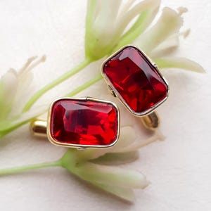 May include: A pair of gold-toned cufflinks with large, rectangular, ruby-red gemstones. The cufflinks are set against a backdrop of white flowers and green stems, creating a striking contrast.