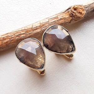 May include: A pair of cufflinks featuring faceted, teardrop-shaped brown gemstones set in gold-toned metal frames. The stones exhibit a translucent, smoky appearance with natural inclusions, and the cufflinks are positioned near a textured wooden branch.