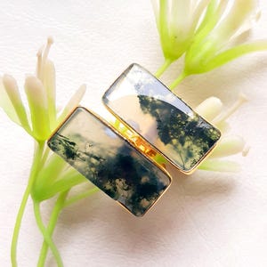 Puede incluir: Dos anillos dorados con piedras rectangulares de ágata musgosa. Las piedras presentan patrones verdes y blancos que se asemejan a paisajes. Los anillos se exhiben sobre una superficie blanca con acentos florales blancos y verdes.