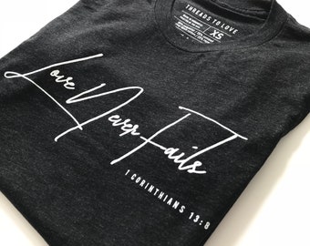May include: A dark grey t-shirt with white lettering that reads "Love Never Fails 1 Corinthians 13:8".