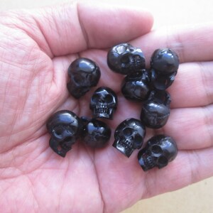 Peut inclure: Un ensemble de perles noires en forme de crâne, probablement en résine ou en plastique, sont éparpillées sur une main. Les perles sont petites et détaillées, avec des orbites et des dents visibles.