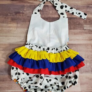 Peut inclure: Un barboteuse blanche pour b&eacute;b&eacute; avec des taches de dalmatien noires, un haut dos nu blanc et une jupe &agrave; volants superpos&eacute;s jaunes, bleus et rouges. La barboteuse a un n&oelig;ud papillon dans le dos avec des taches de dalmatien.