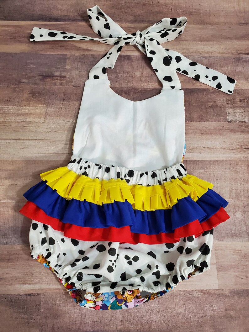 Peut inclure: Un barboteuse blanche pour b&eacute;b&eacute; avec des taches de dalmatien noires, un haut dos nu blanc et une jupe &agrave; volants superpos&eacute;s jaunes, bleus et rouges. La barboteuse a un n&oelig;ud papillon dans le dos avec des taches de dalmatien.