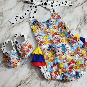 Peut inclure: Ensemble de v&ecirc;tements pour b&eacute;b&eacute; comprenant un bloomer bleu et des chaussures assorties. Le bloomer et les chaussures sont d&eacute;cor&eacute;s d'un motif de chien de dessin anim&eacute; color&eacute;. Le bloomer a un n&oelig;ud et une bordure &agrave; pois blancs et noirs. L'ensemble est con&ccedil;u pour les nourrissons.