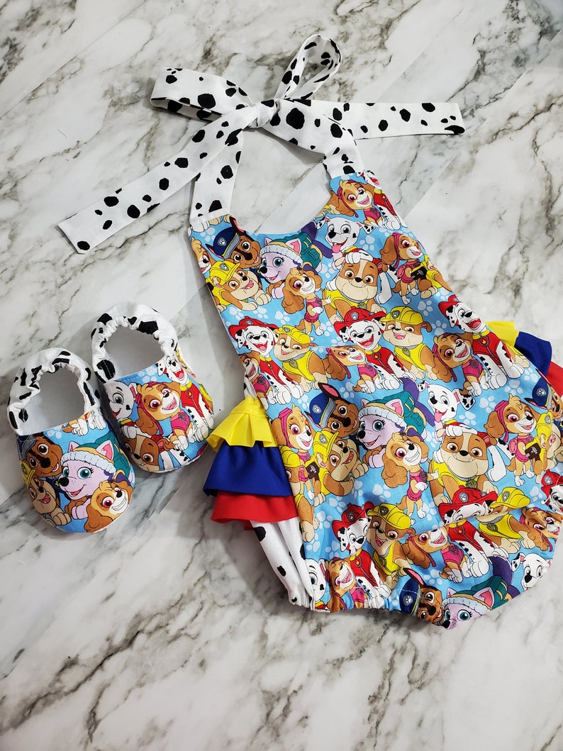 Peut inclure: Ensemble de v&ecirc;tements pour b&eacute;b&eacute; comprenant un bloomer bleu et des chaussures assorties. Le bloomer et les chaussures sont d&eacute;cor&eacute;s d'un motif de chien de dessin anim&eacute; color&eacute;. Le bloomer a un n&oelig;ud et une bordure &agrave; pois blancs et noirs. L'ensemble est con&ccedil;u pour les nourrissons.