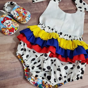 Peut inclure: Ensemble pour b&eacute;b&eacute; comprenant un bloomer blanc &agrave; pois noirs, des volants superpos&eacute;s bleus, jaunes et rouges, et un lien de cou. Des chaussons assortis avec un motif de personnages de dessins anim&eacute;s color&eacute;s sont &eacute;galement inclus.