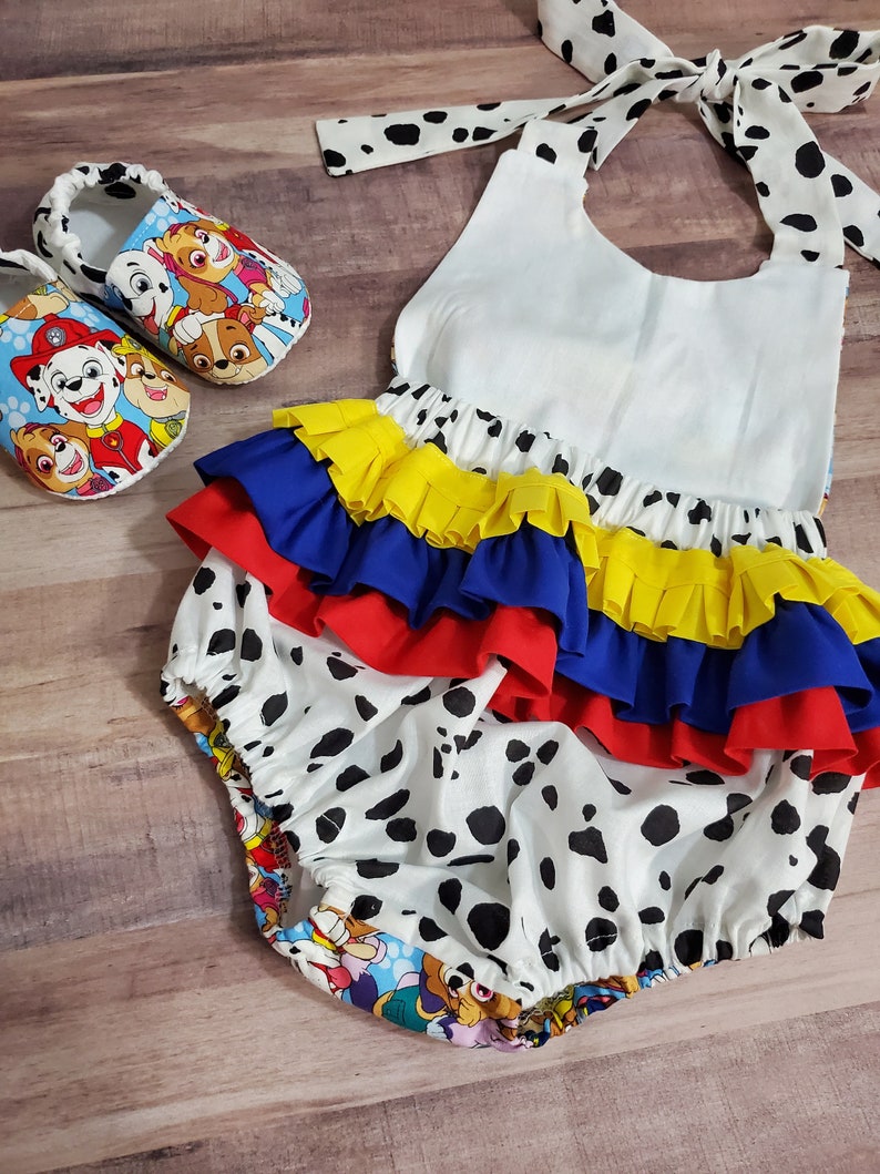 Peut inclure: Ensemble pour b&eacute;b&eacute; comprenant un bloomer blanc &agrave; pois noirs, des volants superpos&eacute;s bleus, jaunes et rouges, et un lien de cou. Des chaussons assortis avec un motif de personnages de dessins anim&eacute;s color&eacute;s sont &eacute;galement inclus.