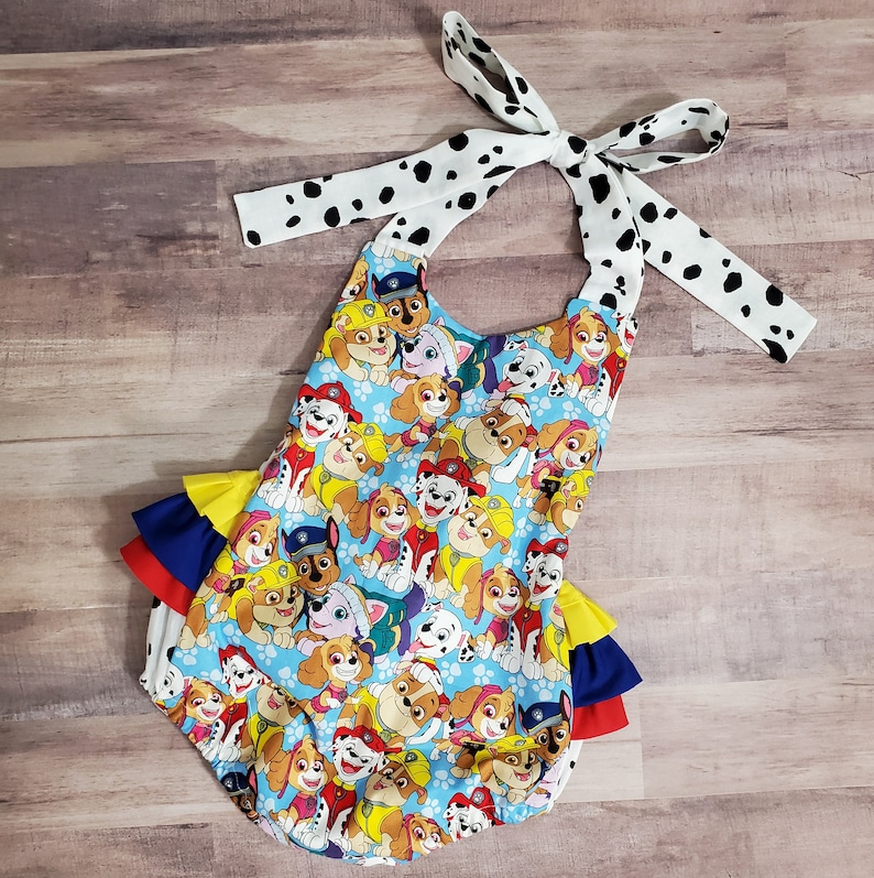 Peut inclure: Un body pour b&eacute;b&eacute; avec un imprim&eacute; de chiots de dessins anim&eacute;s sur fond bleu clair. Il comporte des volants jaunes, bleus et rouges, et est nou&eacute; avec un ruban blanc &agrave; pois noirs.