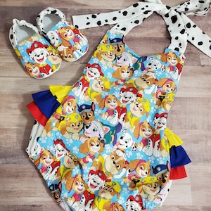 Peut inclure: Ensemble pour b&eacute;b&eacute; comprenant un body bleu et des chaussures assorties, tous deux orn&eacute;s d'un imprim&eacute; Paw Patrol color&eacute;. Le body a des volants superpos&eacute;s bleus, jaunes et rouges. L'ensemble comprend un bandeau blanc &agrave; pois noirs.