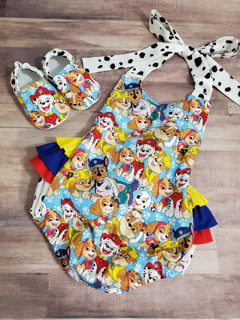 Peut inclure: Ensemble pour b&eacute;b&eacute; comprenant un body bleu et des chaussures assorties, tous deux orn&eacute;s d'un imprim&eacute; Paw Patrol color&eacute;. Le body a des volants superpos&eacute;s bleus, jaunes et rouges. L'ensemble comprend un bandeau blanc &agrave; pois noirs.
