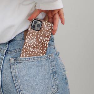 May include: A brown and white spotted phone case is partially visible in a blue jean pocket. The case has a watercolor-like pattern of white spots on a brown background. The phone case is held by a person wearing a white long-sleeved shirt.