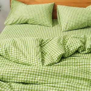 Peut inclure: Une parure de lit avec un motif à carreaux vichy vert clair et blanc. L'ensemble comprend deux taies d'oreiller rectangulaires et une housse de couette assortie drapée sur un lit avec une tête de lit en bois, créant un aspect quadrillé uniforme.