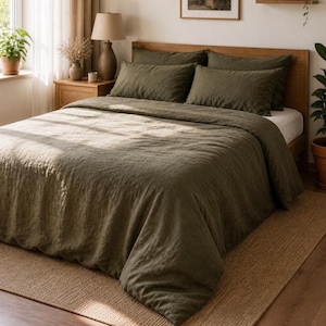 May include: An olive green linen bedding set on a wooden bed frame. The set includes a duvet cover and pillowcases. The bed is placed on a natural fiber rug. A lamp and potted plants are visible in the background, creating a cozy bedroom setting.