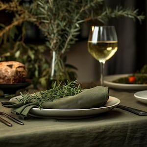 Pode incluir: Um close-up de uma toalha de mesa de linho verde com um prato branco e um guardanapo verde. O guardanapo &eacute; decorado com ramos de alecrim fresco. Um copo de vinho branco e um prato de comida s&atilde;o vis&iacute;veis no fundo.