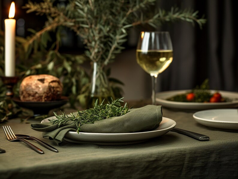 Pode incluir: Um close-up de uma toalha de mesa de linho verde com um prato branco e um guardanapo verde. O guardanapo &eacute; decorado com ramos de alecrim fresco. Um copo de vinho branco e um prato de comida s&atilde;o vis&iacute;veis no fundo.