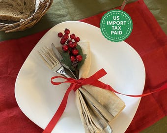 May include: A festive table setting with a white square plate, silverware, and a folded napkin tied with a red ribbon. The napkin is adorned with red berries and greenery. A basket of bread and lit candles are in the background.