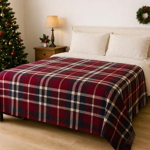 Puede incluir: Una manta rectangular con un patrón de cuadros en rojo oscuro, azul marino y blanco crema. La manta tiene una textura suave, está colocada sobre una cama y muestra un diseño tejido clásico con flecos a lo largo del borde inferior.