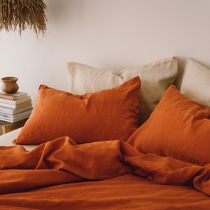 May include: A bed with burnt orange linen bedding and two burnt orange pillowcases. Two cream-colored pillowcases are visible behind the orange ones.