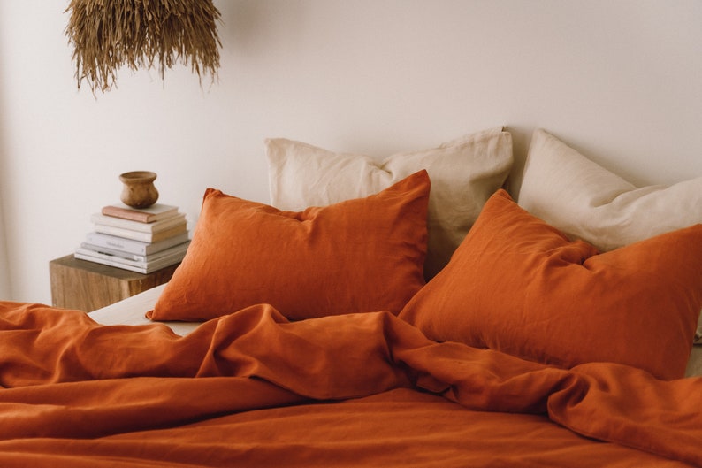 May include: A bed with burnt orange linen bedding and two burnt orange pillowcases. Two cream-colored pillowcases are visible behind the orange ones.