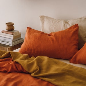 May include: A bed with a rust-colored duvet cover and a mustard yellow throw blanket. There are three pillows on the bed, two are rust-colored and one is cream-colored.