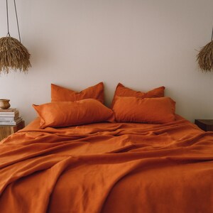 May include: A burnt orange linen bedding set with a duvet cover, two pillowcases, and two shams. The bedding is arranged on a bed with a wooden nightstand on each side. The nightstands have a small lamp with a woven shade hanging above each one.
