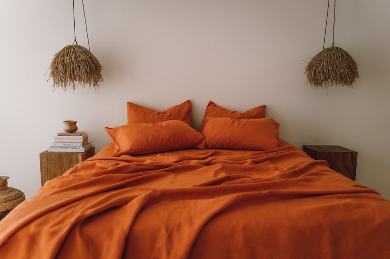 May include: A burnt orange linen bedding set with a duvet cover, two pillowcases, and two shams. The bedding is arranged on a bed with a wooden nightstand on each side. The nightstands have a small lamp with a woven shade hanging above each one.