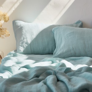 Könnte beinhalten: Ein Bett mit hellblauer Bettwäsche aus Leinen. Die Bettwäsche ist aus einem weichen, natürlichen Stoff gefertigt und hat einen entspannten, lässigen Look. Das Bett ist nicht bezogen, die Laken und Kissen sind zerknittert und leicht schief.