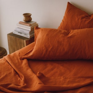 May include: A close-up of a rust-colored linen bedding set with a pillow and a folded sheet. The bedding is arranged on a wooden nightstand with a stack of books and a small wooden bowl.