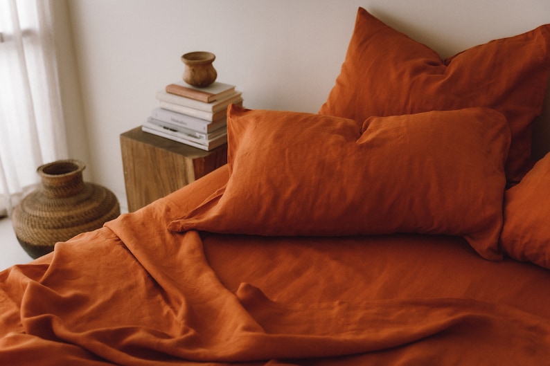 May include: A close-up of a rust-colored linen bedding set with a pillow and a folded sheet. The bedding is arranged on a wooden nightstand with a stack of books and a small wooden bowl.