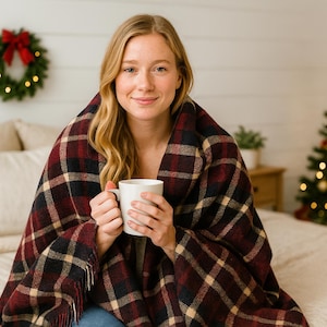 May include: A woman wrapped in a red, black, and tan plaid blanket, holding a white mug. A Christmas tree with lights and ornaments is in the background, along with a wreath. The scene evokes a cozy, festive atmosphere.