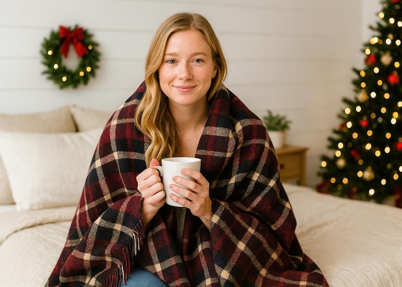 May include: A woman wrapped in a red, black, and tan plaid blanket, holding a white mug. A Christmas tree with lights and ornaments is in the background, along with a wreath. The scene evokes a cozy, festive atmosphere.